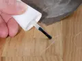 Kleine Kratzer im Parkett reparieren: Ein Finger hält einen Lackstift über eine Holzoberfläche, daneben liegt Schleifpapier.