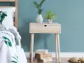 Ein Nachttisch DIY-Projekt aus hellem Holz mit einer Schublade, dekoriert mit Pflanzen und Büchern, neben einem Bett mit Blattmuster.