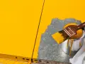 Gelbe Farbe tropft von einem Pinsel, der über eine Dose mit leuchtend gelbem Lack gehalten wird. Ein Teil einer gelben Oberfläche ist sichtbar, während daneben eine graue Fläche mit Farbe bedeckt wird.