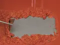 Mit einem Spachtel wird alter, oranger Lack von einer grauen Oberfläche entfernt.