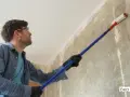 Mann mit Schutzbrille und Handschuhen trägt mit einer Teleskopstange eine Grundierung auf eine Wand auf.