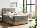 Gemütliches Boxspringbett ohne Lattenrost mit grauem Stoffbezug und Holzsockel. Einladend mit Decken und Kissen dekoriert, bereit für erholsame Nächte.