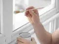 Hand streicht mit Pinsel und weißer Farbe einen weißen Kunststoffrahmen. Dose mit Farbe steht bereit.