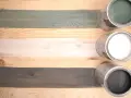 Holzlasur vorher nachher: drei Holzbretter mit unterschiedlichen Lasuren – grün, weiß und grau – und die passenden Dosen.