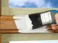 Hand streicht mit weißer Farbe über einen hölzernen Rahmen, um ihn aufzufrischen. Der Spiegelrahmen wird sorgfältig mit Klebeband geschützt.