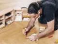 Mann repariert den Holzboden mit einem Reparaturstift und einer Farbkarte. Werkzeugkasten mit Zubehör daneben.