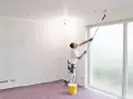 Maler streicht Decke und Wand mit langem Teleskoproller, um das Streichen ohne Abkleben zu ermöglichen. Gelber Farbeimer steht bereit.