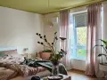 Gemütliches Schlafzimmer mit Bett in Khaki-Farbe, grüner Decke und rosa Vorhängen. Ein Blumenstrauß steht auf einem Holztisch.