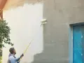 Mann streicht mit einer Teleskoprolle die Hausfassade weiß. Die rechte Seite ist noch grau, die linke bereits frisch gestrichen.