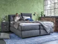 Frau entspannt auf einem Boxspringbett mit grauem Stoffbezug. Das Bett bietet hohen Komfort, was ein Boxspringbett ausmacht.