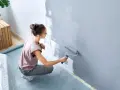 Frau streicht Wand grau. Nützliche **Wand streichen Tipps** für ein gleichmäßiges Ergebnis. Möbeltisch mit Folie abgedeckt.