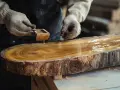 Hände mit Handschuhen tragen goldenes Öl auf einen rustikalen Baumscheiben-Tisch auf, um das Holz zu lasieren und seine natürliche Schönheit hervorzuheben.