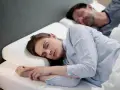 Frau schläft friedlich auf einem Nackenkissen, das zeigt, wie man ein Nackenkissen richtig benutzt. Mann schläft im Hintergrund.