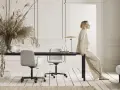 Eine Frau in einem beigen Anzug sitzt auf einem Schreibtisch. Der Raum ist in neutralen Farben gehalten, mit einem großen Trockenblumenstrauß und zwei Bürostühlen.