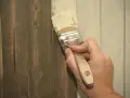 Hand streicht mit Pinsel über dunkles Holz, um es aufzuhellen. Ein Teil der Wand ist bereits heller gestrichen.