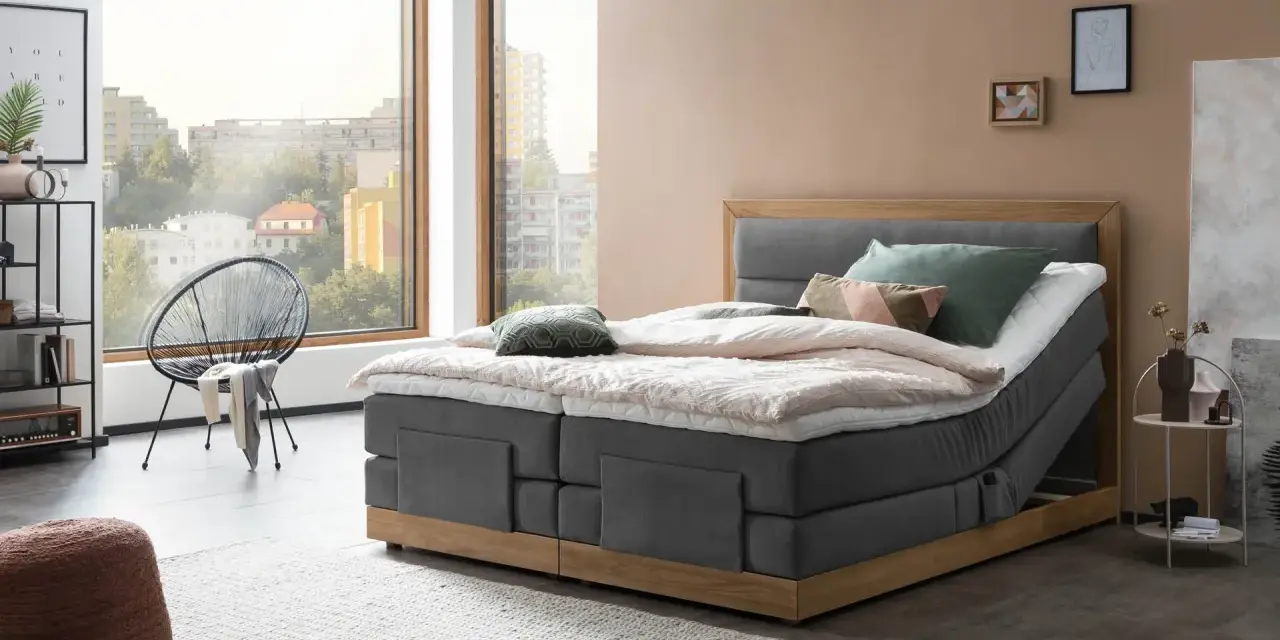 Modernes Boxspringbett mit Holzrahmen und grauem Stoffbezug. Die Matratze wirkt etwas zu hart, aber die Kissen laden zum Entspannen ein.