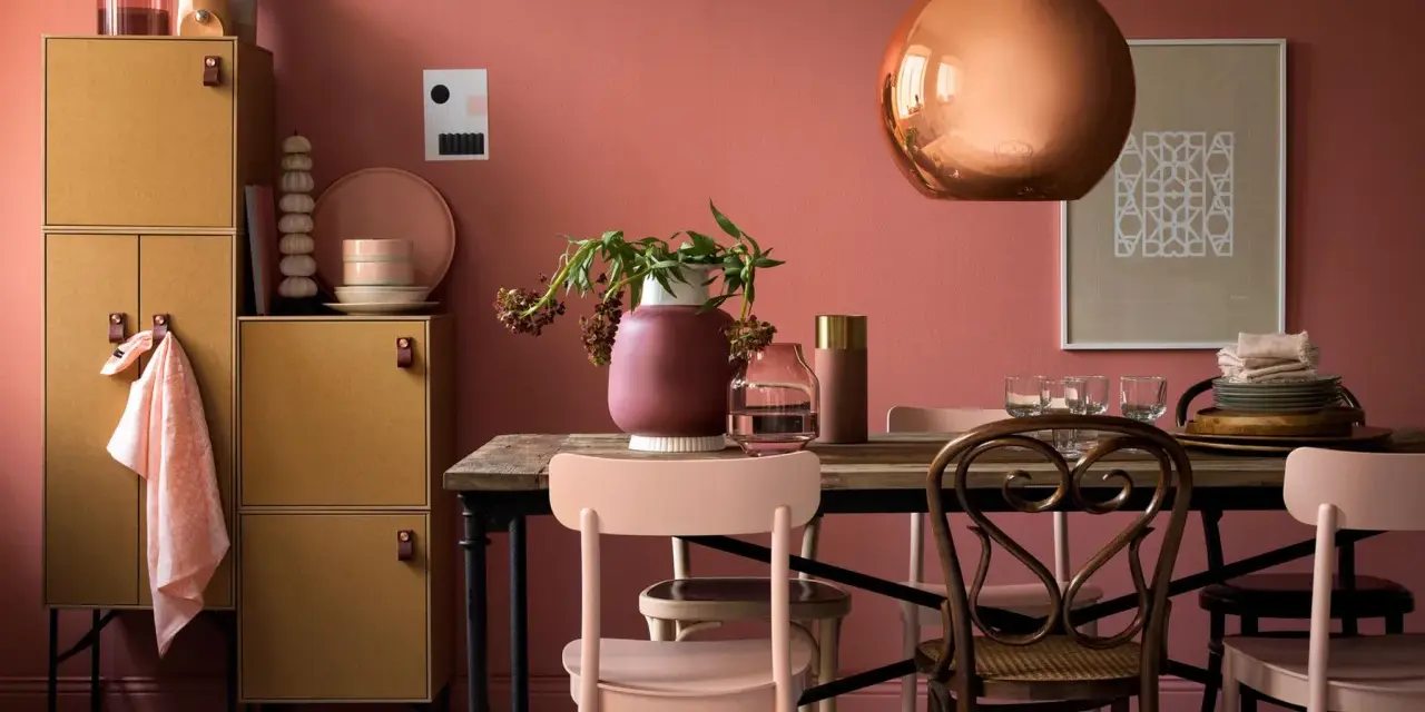 Ein Esszimmer mit altrosa Wänden. Ein kupferfarbener Lampenschirm und ein Holzmöbelstück mit gelben Türen. Welche Farbe zu altrosa? Zarte Rosatöne und Erdtöne passen gut.