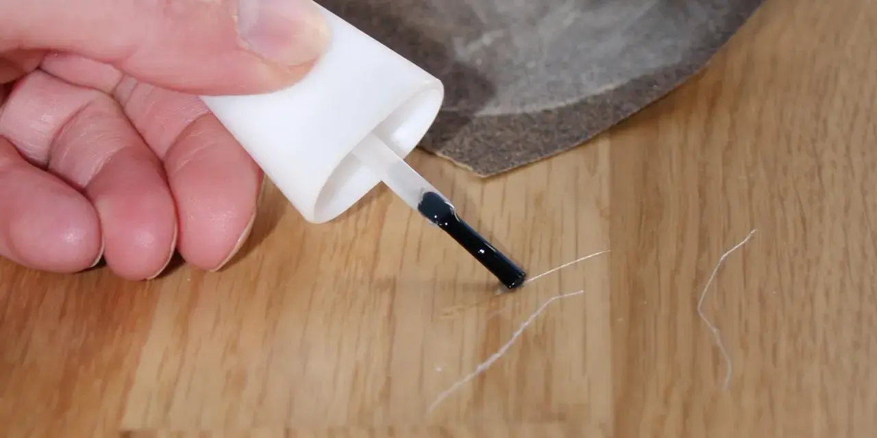 Kleine Kratzer im Parkett reparieren: Ein Finger hält einen Lackstift über eine Holzoberfläche, daneben liegt Schleifpapier.