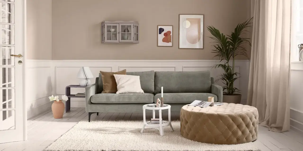 Ein Wohnzimmer mit salbeigrünem Sofa. Braune und cremefarbene Kissen, ein beiger Hocker und eine helle Wand zeigen, welche Farbe passt zu Salbei.