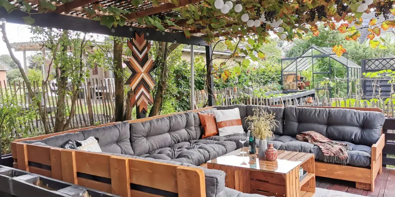Gemütliche Paletten Lounge Ideen für den Garten. Ein großer Ecksofa aus Paletten mit grauen Polstern und Holztisch. Weinreben und Lichterketten überdachen den Sitzbereich.