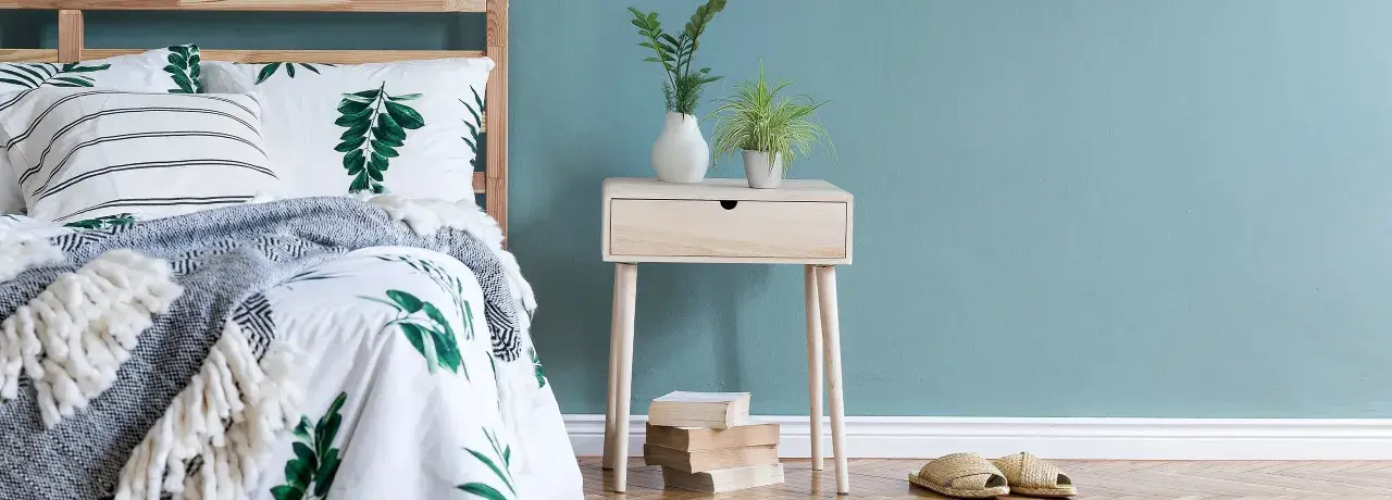 Ein Nachttisch DIY-Projekt aus hellem Holz mit einer Schublade, dekoriert mit Pflanzen und Büchern, neben einem Bett mit Blattmuster.