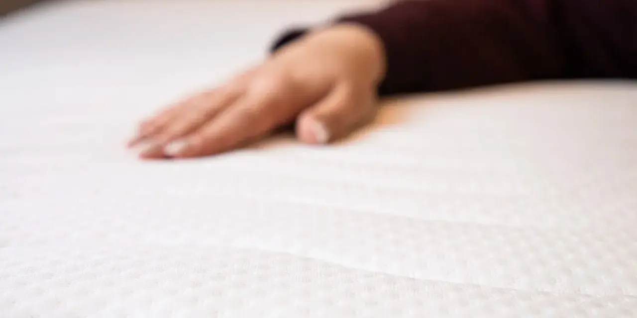 Eine Hand liegt auf einer weißen Matratze. Wann sollte man eine Matratze austauschen? Wenn sie durchgelegen ist oder Flecken hat.