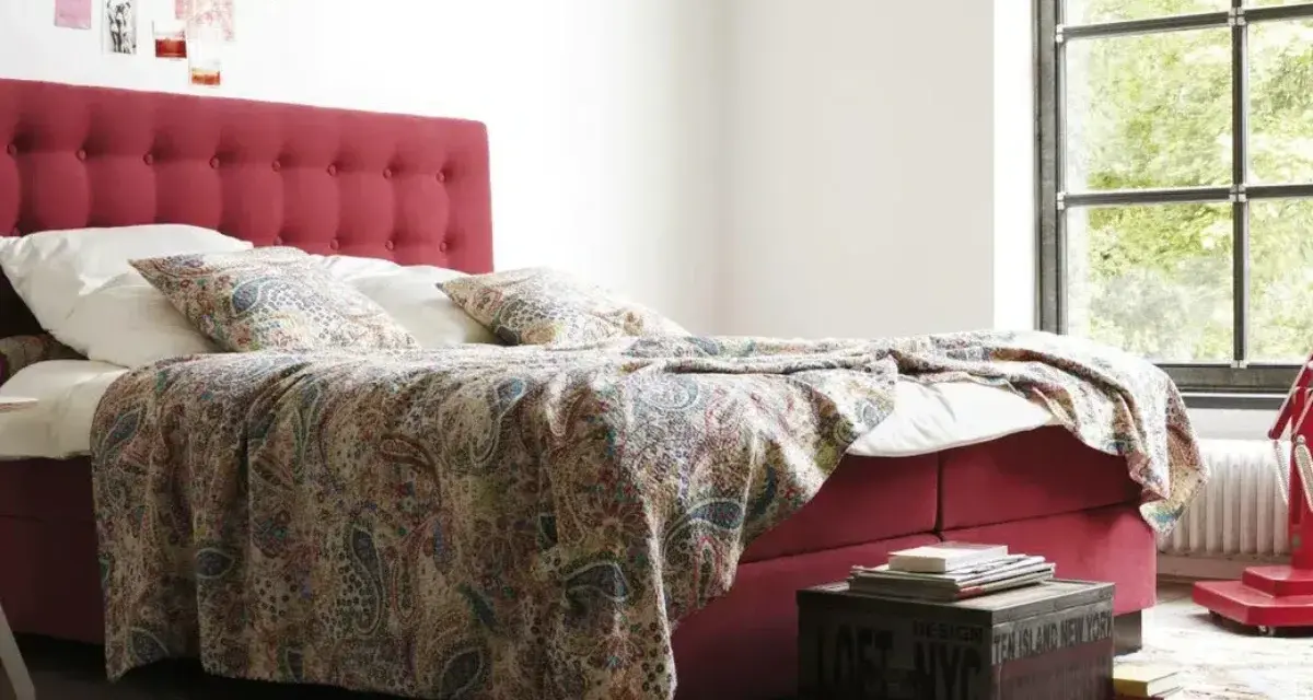 Rotes Boxspringbett mit gemustertem Überwurf und Kissen, ideal für Allergiker. Ein Nachttisch mit Büchern steht daneben.