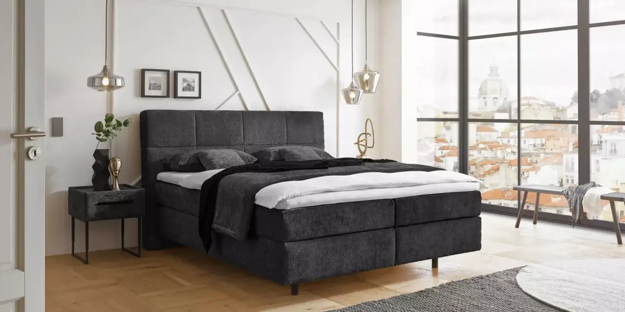 Ein modernes Schlafzimmer mit einem dunklen Boxspringbett ja oder nein? Helle Wände, Holzboden und ein Fenster mit Stadtblick.