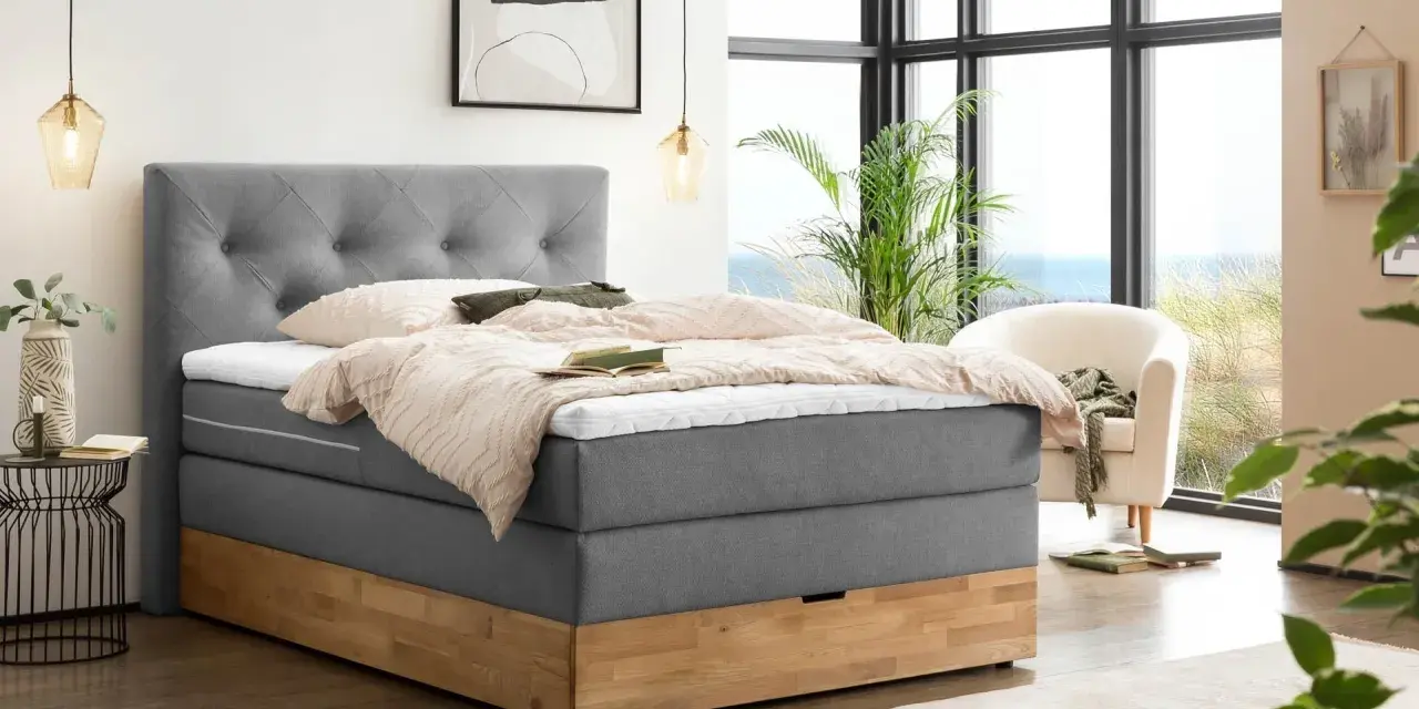 Gemütliches Boxspringbett ohne Lattenrost mit grauem Stoffbezug und Holzsockel. Einladend mit Decken und Kissen dekoriert, bereit für erholsame Nächte.