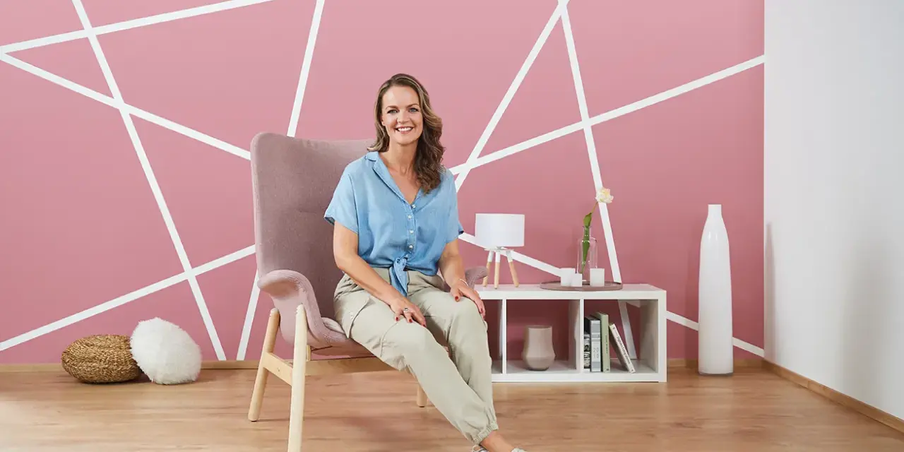 Frau sitzt vor einer rosa Wand, die mit weißen Linien diagonal gestrichen ist.