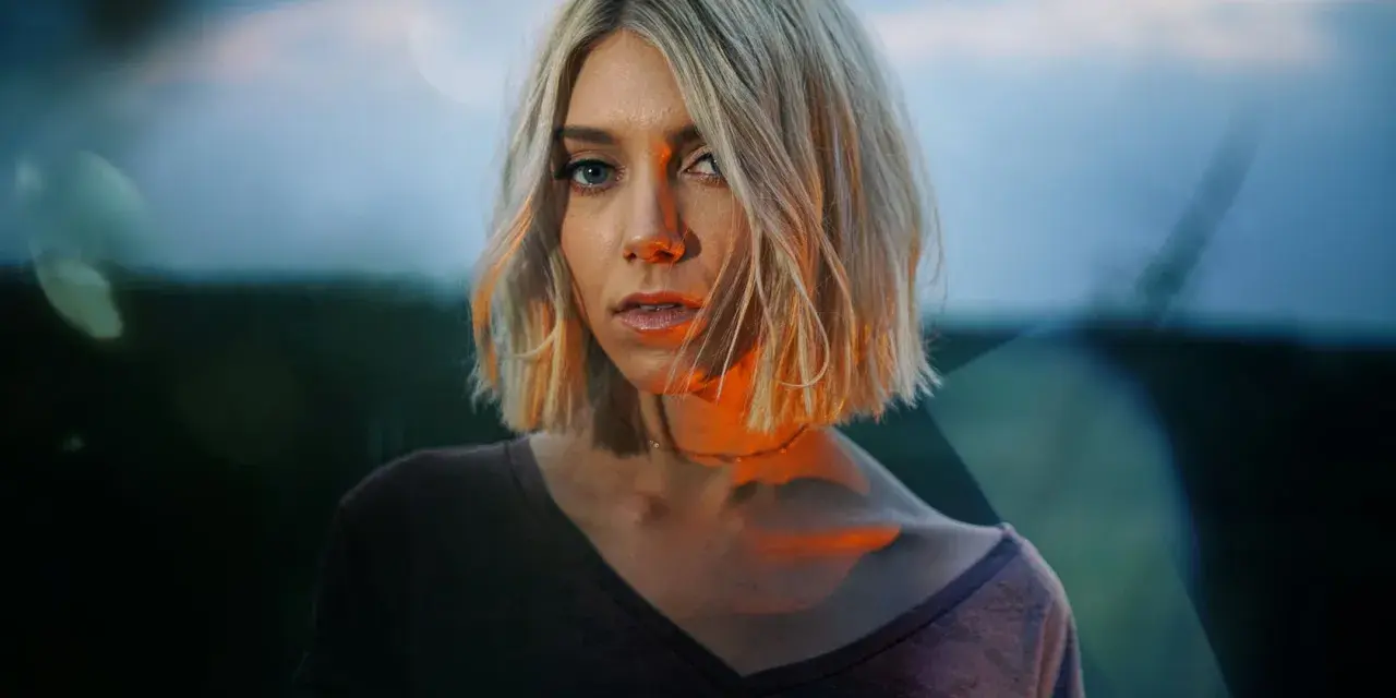 Blonde Frau mit Bob-Schnitt, deren Gesicht von warmem Licht beleuchtet wird, das sich mit dem blauen Abendhimmel mischt.