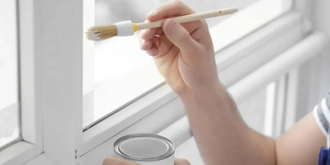 Hand streicht mit Pinsel und weißer Farbe einen weißen Kunststoffrahmen. Dose mit Farbe steht bereit.