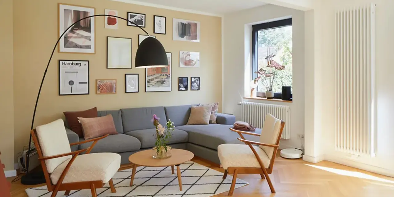Gemütliches Wohnzimmer mit grauem Sofa, zwei Sesseln und vielen Bildern an der gelben Wand. Perfekt, um die Wand abzukleben und neu zu gestalten.