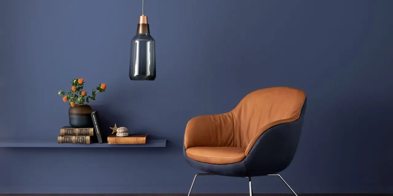 Ein Sessel mit braunem Leder und dunkelblauem Stoff vor einer blauen Wand. Orange Blumen auf einem Regal zeigen, welche Farbe passt zu Blau.