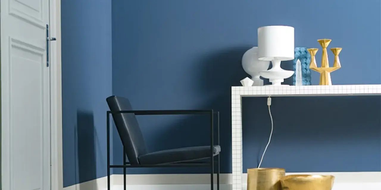 Ein Sessel vor einer Wand in moderner Wandfarbe. Auf einem Regal stehen eine Lampe, eine Vase und ein goldener Kerzenständer.