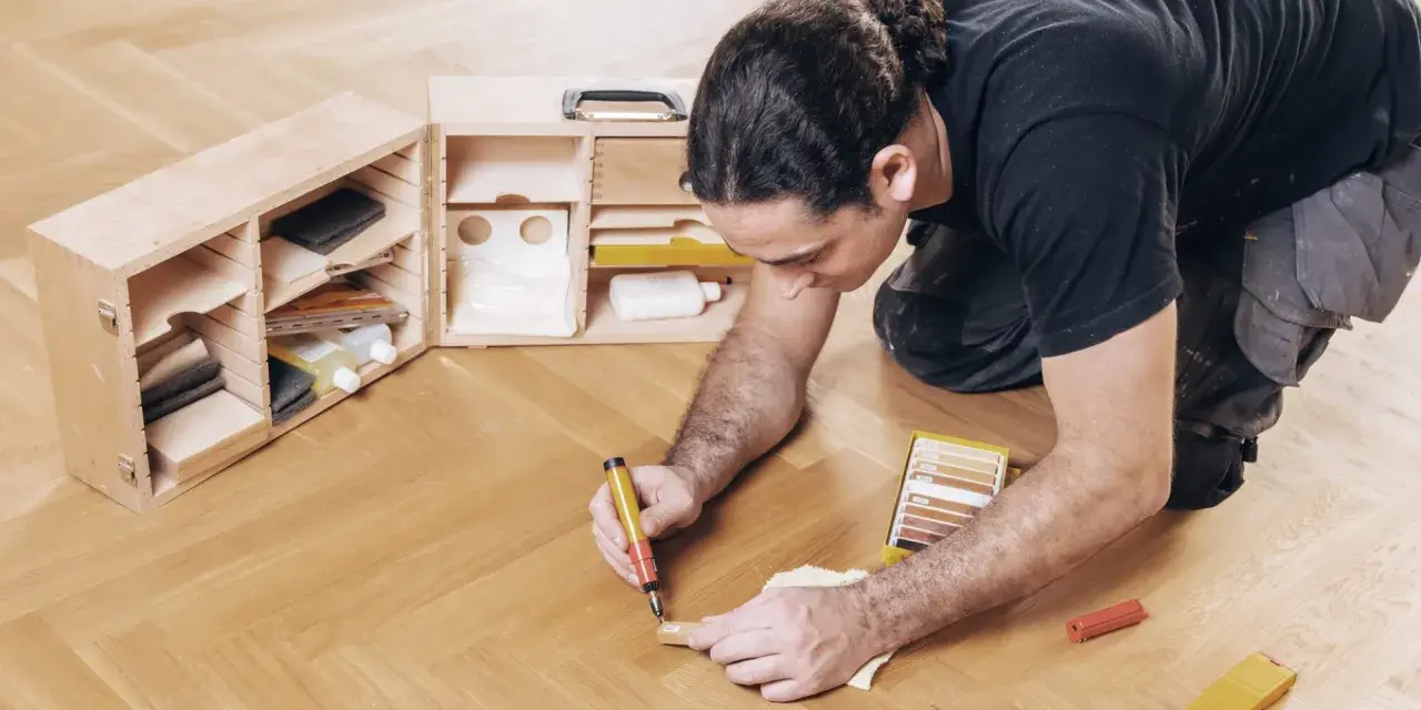 Mann repariert den Holzboden mit einem Reparaturstift und einer Farbkarte. Werkzeugkasten mit Zubehör daneben.