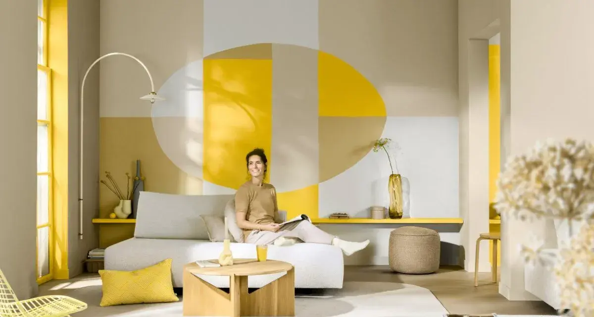 Eine Frau sitzt auf einem Sofa in einem hellen Wohnzimmer. Die Wand hinter ihr ist mit geometrischen Formen in Gelb, Beige und Grau gestaltet. Die Frage, welche Farbe passt zu Gelb, wird hier mit warmen Erdtönen und neutralen Farben beantwortet.