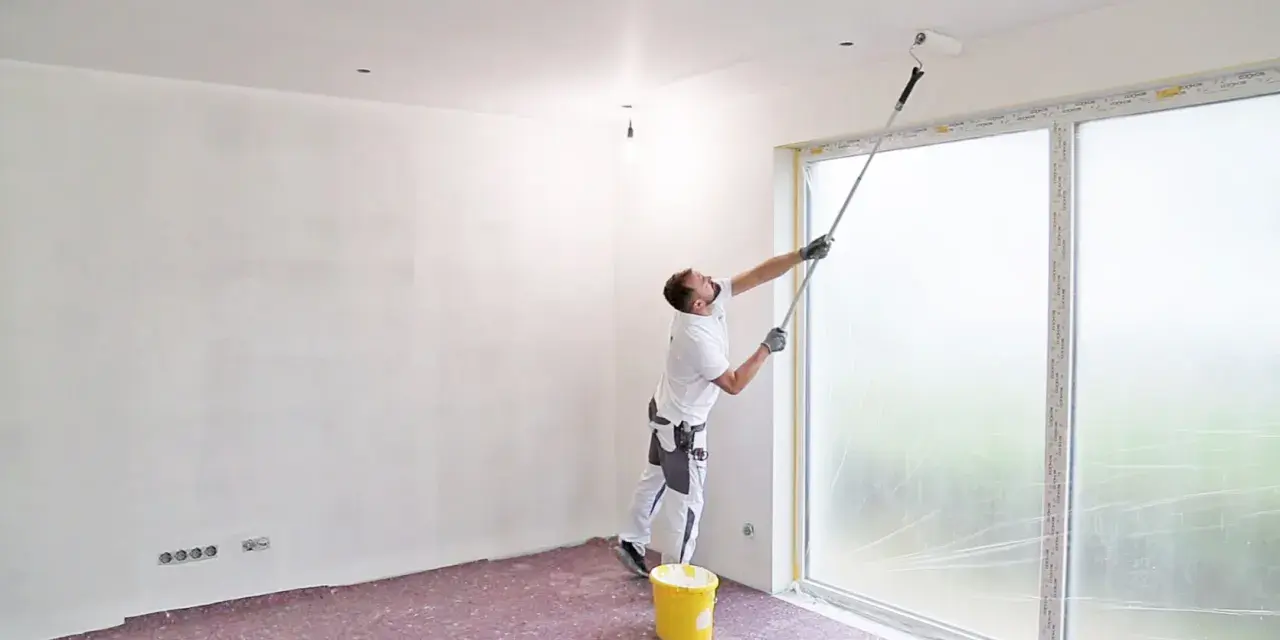 Maler streicht Decke und Wand mit langem Teleskoproller, um das Streichen ohne Abkleben zu ermöglichen. Gelber Farbeimer steht bereit.