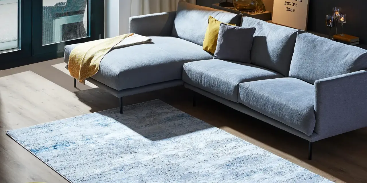 Ein moderner Wohnbereich mit einem grauen Ecksofa und einem Teppich, der richtig platziert unter dem Sofa liegt.