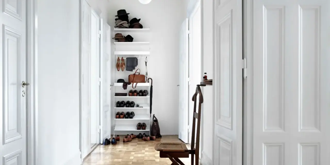 Schmaler Flur mit weißer Wandfarbe, Holzboden und einem Regal voller Schuhe und Hüte. Ein Stuhl steht bereit.