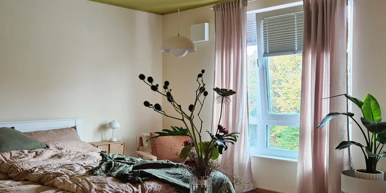 Gemütliches Schlafzimmer mit Bett in Khaki-Farbe, grüner Decke und rosa Vorhängen. Ein Blumenstrauß steht auf einem Holztisch.