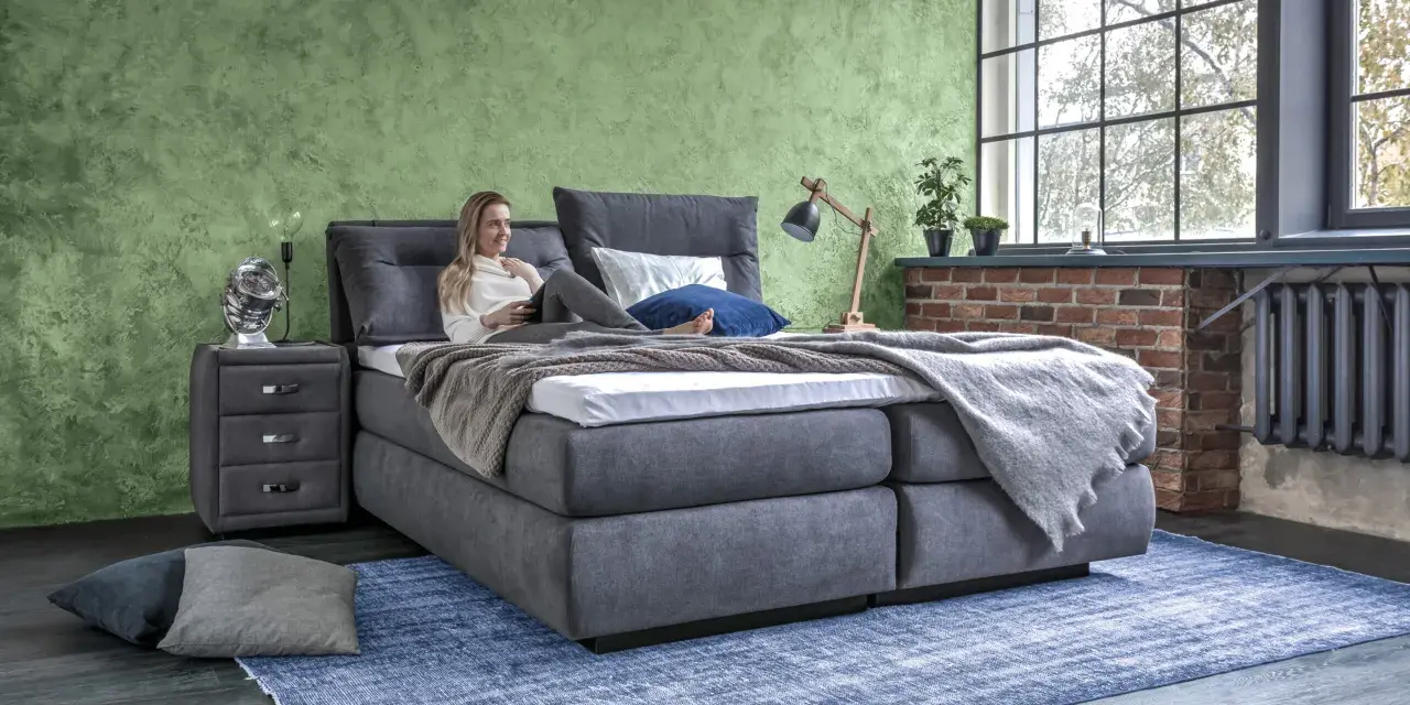 Frau entspannt auf einem Boxspringbett mit grauem Stoffbezug. Das Bett bietet hohen Komfort, was ein Boxspringbett ausmacht.