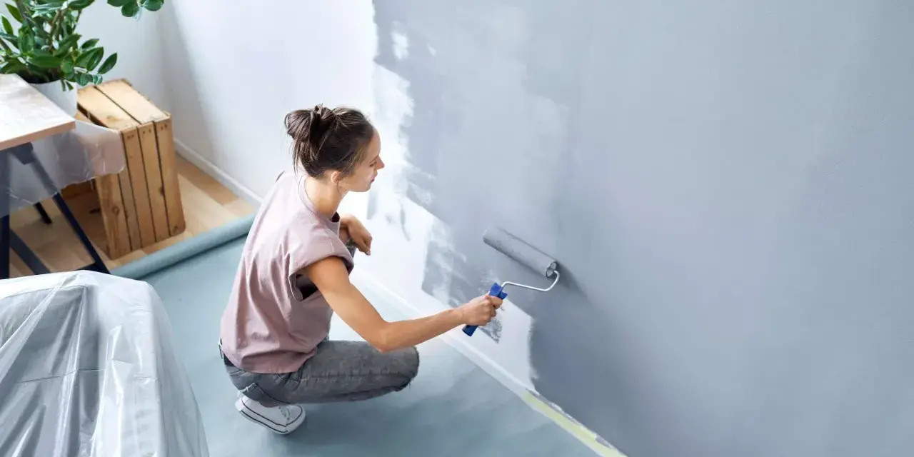 Frau streicht Wand grau. Nützliche **Wand streichen Tipps** für ein gleichmäßiges Ergebnis. Möbeltisch mit Folie abgedeckt.
