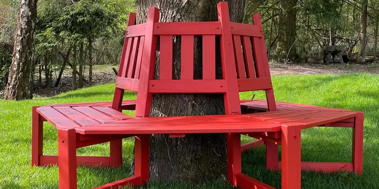 Frisch gestrichene rote Gartenbank um einen Baum im Grünen.