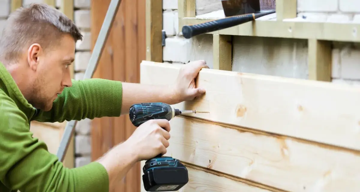 Mann renoviert Holzfassade, schraubt neue Bretter an. Werkzeug liegt bereit.