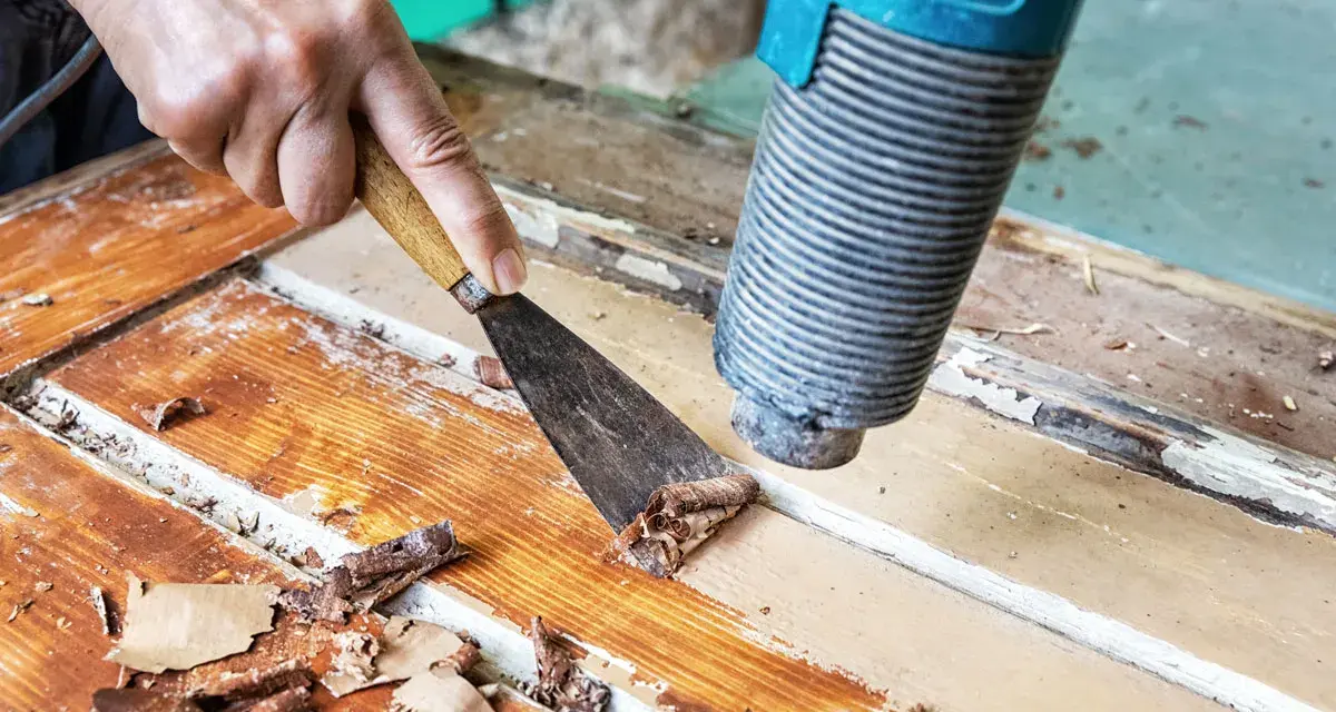 Farbe von Holz entfernen ohne Schleifen: Heißluftföhn und Spachtel lösen alte Lackschichten von einer Holztür.