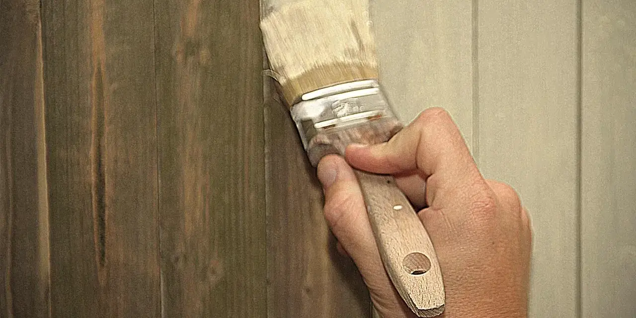 Hand streicht mit Pinsel über dunkles Holz, um es aufzuhellen. Ein Teil der Wand ist bereits heller gestrichen.