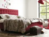 Rotes Boxspringbett mit gemustertem Überwurf und Kissen, ideal für Allergiker. Ein Nachttisch mit Büchern steht daneben.