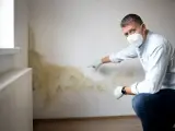 Mann mit Maske und Handschuhen zeigt auf schimmeligen Putz an einer Wand neben einem Heizkörper.