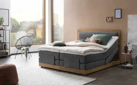 Modernes Boxspringbett mit Holzrahmen und grauem Stoffbezug. Die Matratze wirkt etwas zu hart, aber die Kissen laden zum Entspannen ein.